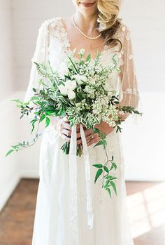 a woman in a white wedding dress holding a bouquet with greenery and flowers on it