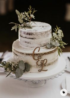 a three tiered wedding cake with greenery and initials on the top is sitting on a table