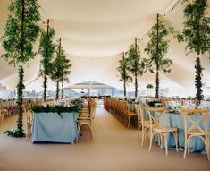 an indoor tent with tables and chairs covered in blue linens, trees and greenery