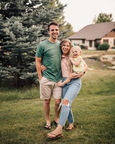 a man and woman are standing in the grass with a baby on their lap, smiling at the camera