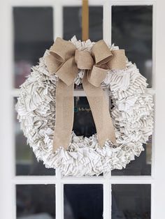 a white wreath with a tan bow hangs on the front door window sill, decorated with jute and burlap