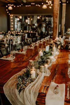 a long wooden table topped with lots of candles and place settings covered in greenery