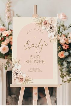 a welcome sign for a baby shower with pink flowers and greenery on it in front of a fireplace