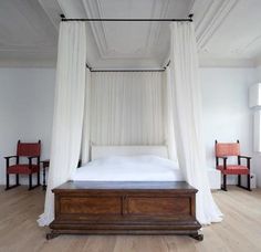 a canopy bed with white drapes over it in a room filled with wooden floors and red chairs