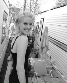 black and white photograph of a woman standing next to a camper trailer smiling at the camera