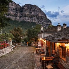 a man sitting at a table in the middle of a cobblestone street with mountains in the background