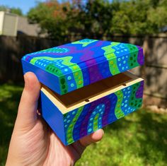 a hand is holding up a colorful box in the grass with trees in the background