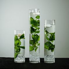 three tall glass vases filled with water and green leaves on the inside, along with a candle