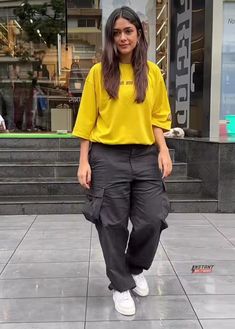 a woman in yellow shirt and black pants standing on the sidewalk with her hands in her pockets