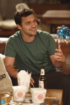 a man sitting on a couch talking to another man who is holding a beer and eating