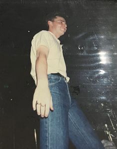 a man in white shirt and blue jeans standing next to a black wall with his hands out