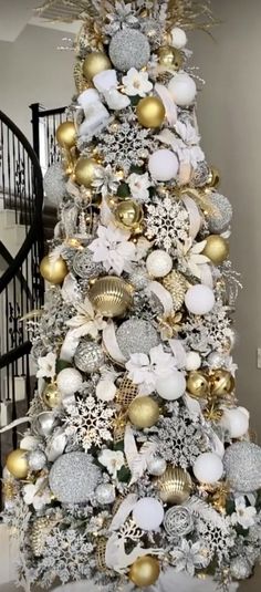 a white christmas tree with gold and silver ornaments on it's top, in front of a spiral staircase