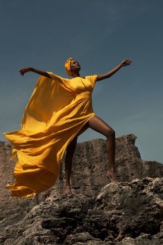 a woman in a yellow dress standing on top of a rocky hill with her arms outstretched