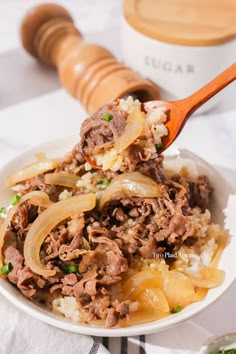 a bowl filled with rice and meat on top of a table next to a wooden spoon