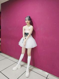 a girl in a tutu and short skirt posing against a pink wall with her hand on her hip