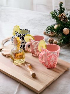 a wooden cutting board topped with two cups filled with liquid and lemon slices next to a pine tree