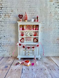 a white china cabinet with pink and red decorations on the top, sitting in front of a brick wall