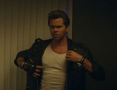 a man in white shirt and black leather jacket holding his hand on his shoulder while standing next to a wall