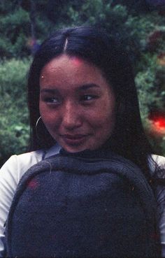 a woman with long black hair wearing a gray backpack and smiling at the camera while standing in front of some bushes