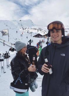 two people standing in the snow with skis on and holding beer bottles, smiling