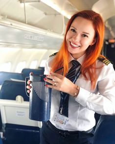 a woman with red hair wearing a tie and holding a cup in an airplane cabin