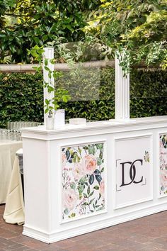 an outdoor bar with flowers and greenery on the front, surrounded by white tablecloths