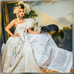 a woman in a white dress and feathered hat standing next to large sheet of paper