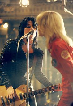 two women singing into microphones while one plays an electric guitar in front of the other