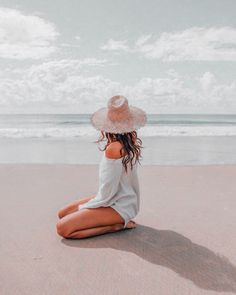 a woman sitting on the beach wearing a hat