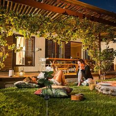 two people sitting on the grass in front of a house with an outdoor dining area