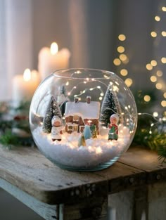 a glass bowl filled with snow and christmas decorations on top of a wooden table next to candles