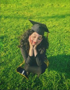 a woman sitting in the grass wearing a graduation cap and holding her hands to her face