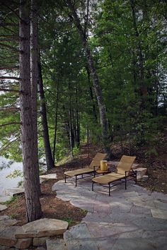 two lounge chairs sitting on top of a stone patio next to a forest filled with trees