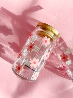 two glasses with straws in them sitting on a pink tablecloth, one is decorated with flowers and the other has a gold lid