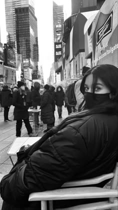 a woman sitting on a bench in the middle of a city street wearing a face mask