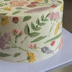 a white cake with flowers and leaves painted on it's side, sitting on a table