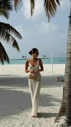 a woman in a white dress standing on the beach next to a palm tree and looking at her cell phone