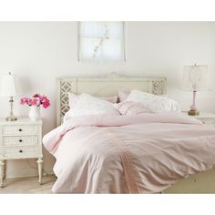 a bed with pink sheets and pillows in a white room next to a table with flowers on it