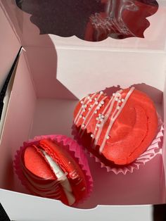 two red cupcakes with white frosting in a pink box on a table