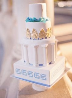 a three tiered white cake with blue and gold decorations on the top, sitting on a table