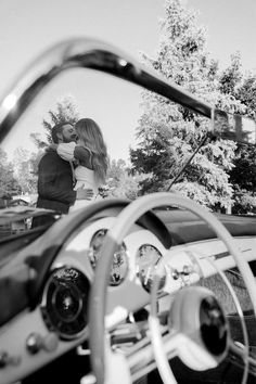 a man and woman kissing in the drivers seat of a vintage car with trees in the background