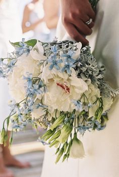 a bride holding a bouquet of white and blue flowers in her hand while standing next to the groom