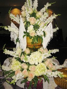 an arrangement of white flowers and greenery in a gold vase with fringes on top