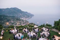 a group of people sitting at tables on top of a lush green hillside next to the ocean