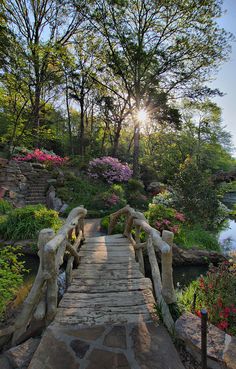 a wooden bridge crossing over a river surrounded by trees and flowers with the sun shining down