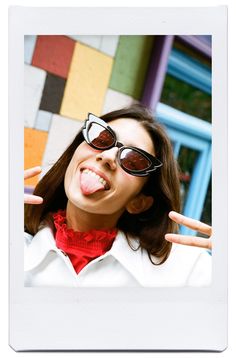 a woman with her tongue out and sunglasses on, making the peace sign in front of a multi - colored building