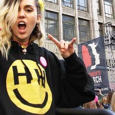 a woman wearing a smiley face sweatshirt and holding her hands up in the air while standing next to a crowd