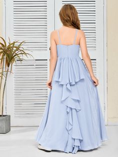 a woman in a long blue dress is standing near a window with shutters and a potted plant