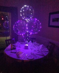 the table is set for dinner with purple lights in the room and balloons floating over it