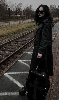 a woman standing on train tracks with her suitcase in front of her, wearing sunglasses and a black coat